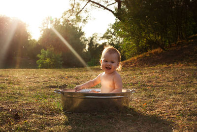 Cute little kid sits in a basin of water in nature and has rays of the setting sun, golden bathe. 