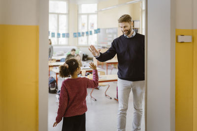 Happy male teacher giving high-five to schoolgirl at doorway in school