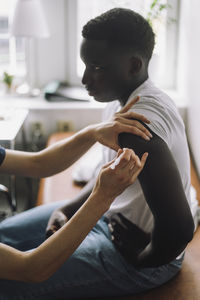 Side view of male teenage patient taking vaccination on arm while sitting in clinic