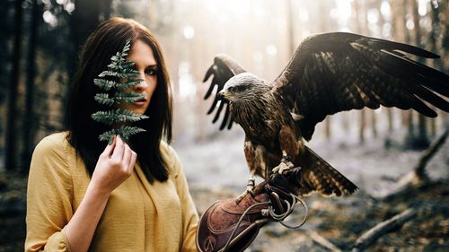Portrait of beautiful woman standing with hawk while holding leaf in forest
