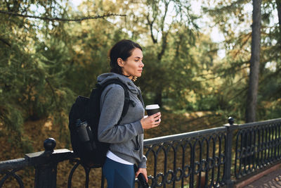 Young woman looking away while standing by railing against trees