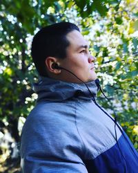 Side view of young man listening to music while standing against trees
