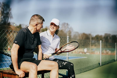Smiling senior friends talking while sitting on bench at tennis court