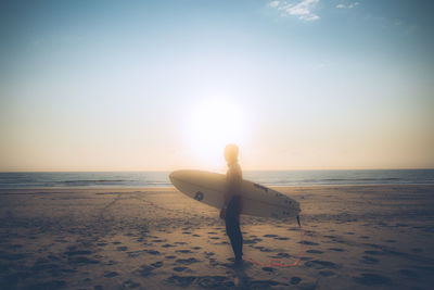 Man carrying surfboard at beach against sky during sunset