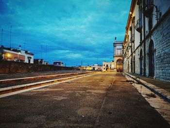 Empty road by illuminated buildings against sky at dusk