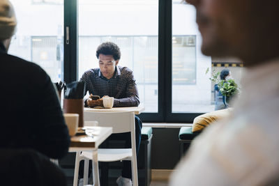 Young customer using mobile phone while sitting with coffee cup at table in cafe