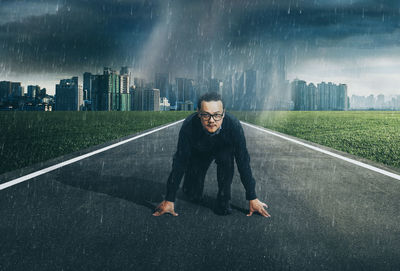 Portrait of businessman kneeling on road against cityscape during rainy season