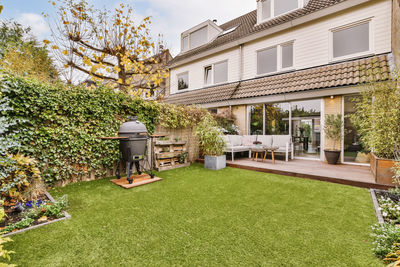 Cozy wooden table and soft sofa and grill placed in backyard with green garden near contemporary residential building