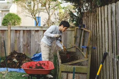 A man uses a pitchfork to shovel compost into a red wheelbarrow