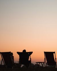 Rear view of people relaxing on deck chairs against clear sky during sunset