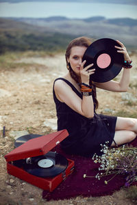 Young woman sit in nature in a black dress next to an old gramophone and listens to music