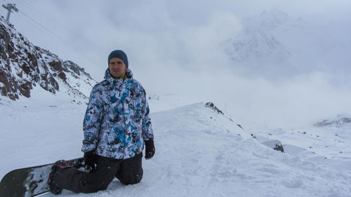 A male snowboarder kneeling on a ski slope with his board strapped in. in the background 