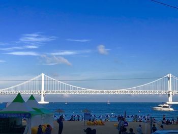 People at beach with suspension bridge over sea in background against blue sky