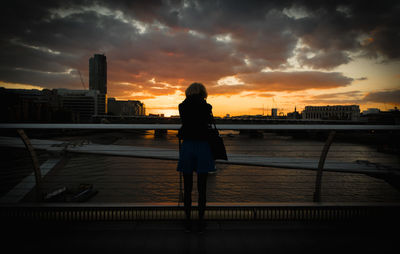 Woman standing on railing by river against sky during sunset