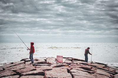 Men fishing by sea against cloudy sky during sunny day