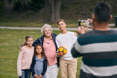 Rear view of man photographing happy grandmother and grandchildren in park during picnic