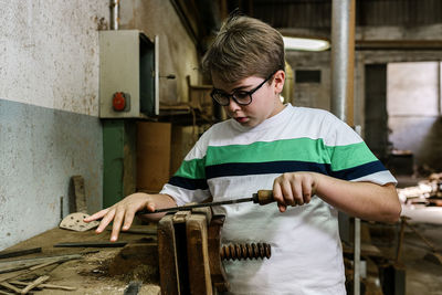 Serious teenage boy grinding piece of wood in vice using metal rasp while working in shabby workshop