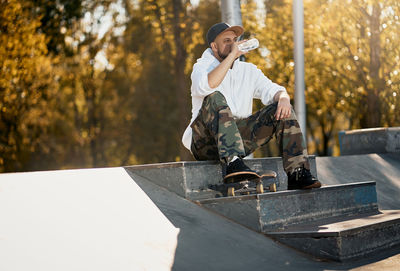 Full length of man with skateboard drinking water while sitting on steps