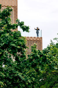 Low angle of female in stylish outfit using vr goggles while standing on terrace of house near glass railings under cloudy sky in daylight near green trees and plants