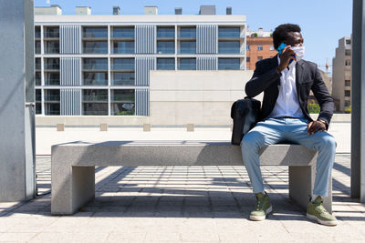 Black male entrepreneur wearing medical mask sitting on stone bench in city and discussing work issues on cellphone during covid 19 epidemic