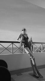 Full length of woman wearing swimsuit leaning on railing in balcony against sky