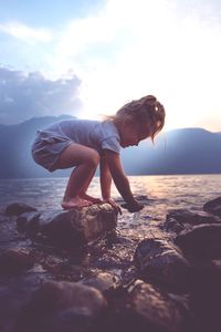 Side view of girl standing on rock at beach against cloudy sky