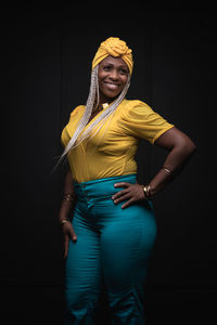 Smiling young african american female with dyed afro braids wearing yellow clothes and jeans with headscarf and necklace on black background