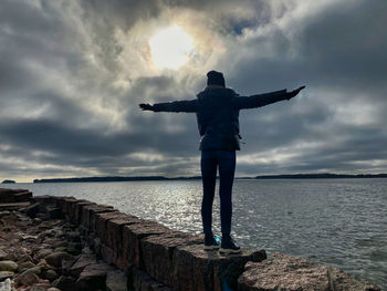 Rear view of woman standing with arms outstretched by sea against sky