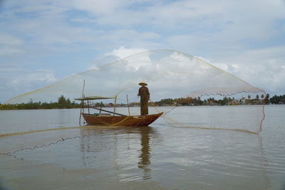 Fisherman throwing net while standing in boat on sea against sky