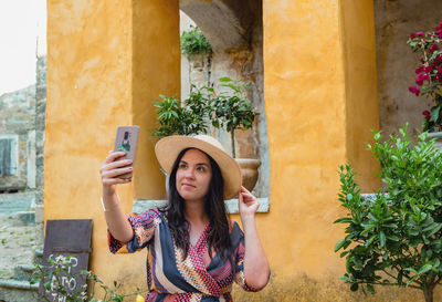 Beautiful young woman using mobile phone, taking a selfie in front of old colorful yellow building