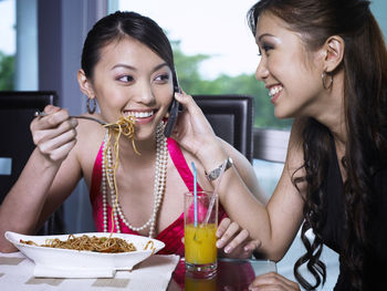 Female friends eating spaghetti while talking on mobile phone in restaurant