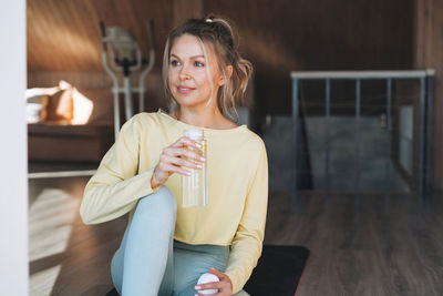 Young fitness blonde woman with bottle of water with lemon doing morning exercises near window 