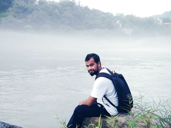 Portrait of young man sitting on rock by lake during foggy weather