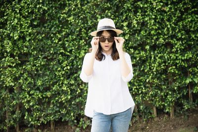 Portrait of young woman wearing hat and sunglasses while standing against plants