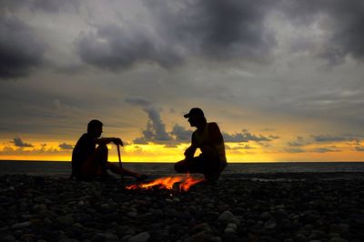 Silhouette friends by campfire at beach against sky during sunset