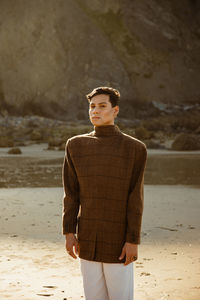 Young ethnic man looking at camera and wearing stylish jacket while relaxing on sandy beach near sea and cliff in summer