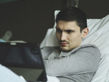A young man with a tablet lies in bed in a hospital.