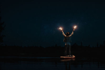 Rear view of woman standing with sparkler on paddleboard in lake at night