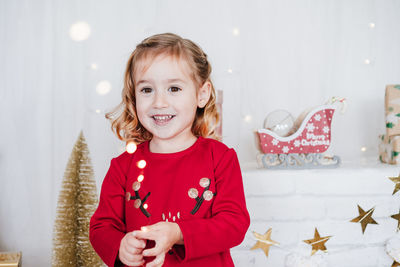 Smiling girl at home holding garland lights wearing red christmas dress at home over christmas deco
