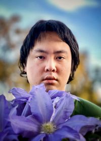 Portrait of young man with purple clematis flowers against sky and trees.