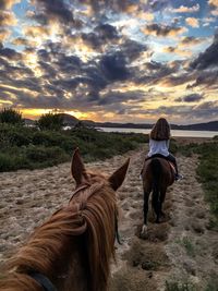 Rear view of woman horseback riding on field against cloudy sky during sunset
