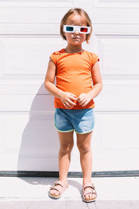 Cute girl in casual colorful clothes and three dimensional glasses standing on white wall background looking at camera