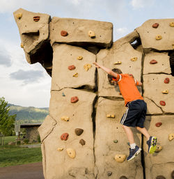 Side view of boy climbing boulder against sky at playground