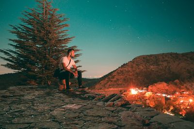 Young man sitting by tree against sky during winter at night