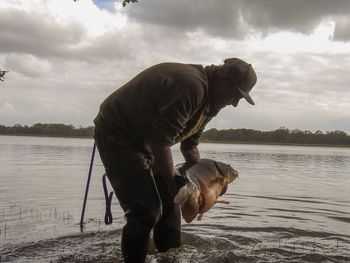 Man holding fish while standing in lake against sky