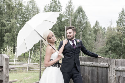 Bride with groom holding umbrella against trees