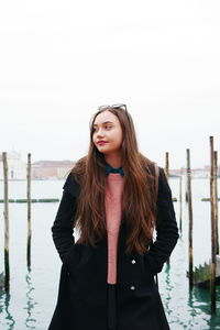 Portrait of beautiful young woman standing at lakeshore against clear sky