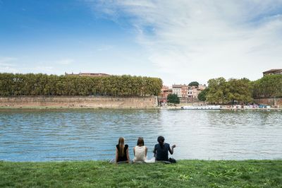 Rear view of women sitting on riverbank against sky