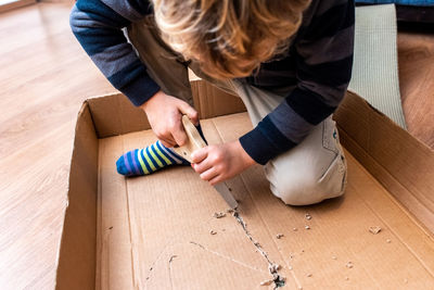 Boy cutting cardboard with work tool while sitting on floor