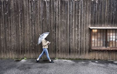 Side view of young woman with umbrella walking on road against wooden wall